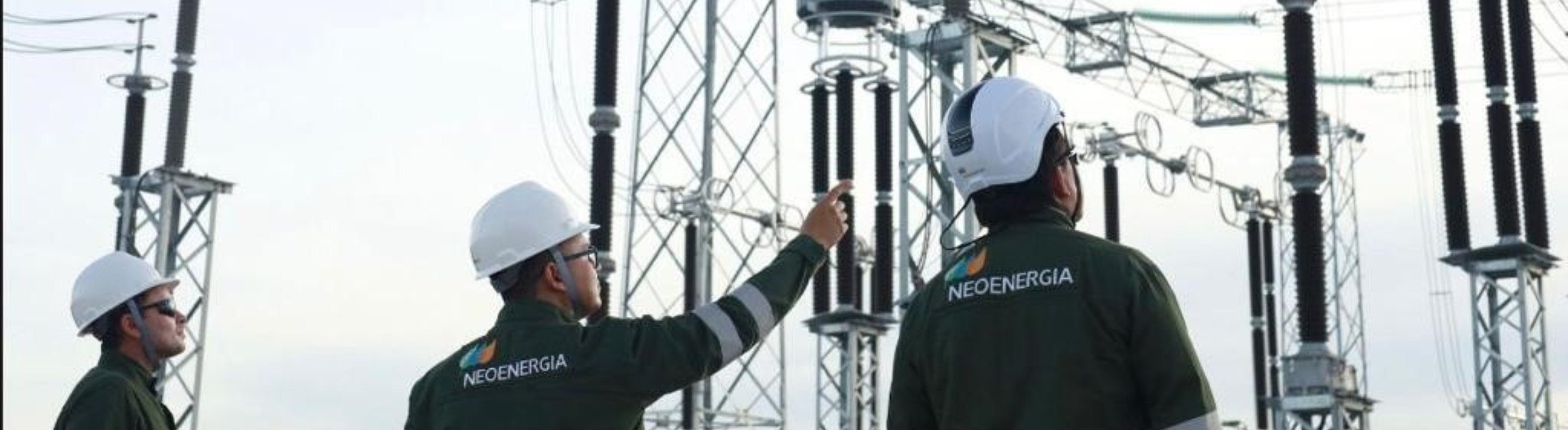 Imagem mostra três homens vestindo macacões verde com a inscrição Neoenergia. Eles usam capacetes brancos e observam instalações elétricas