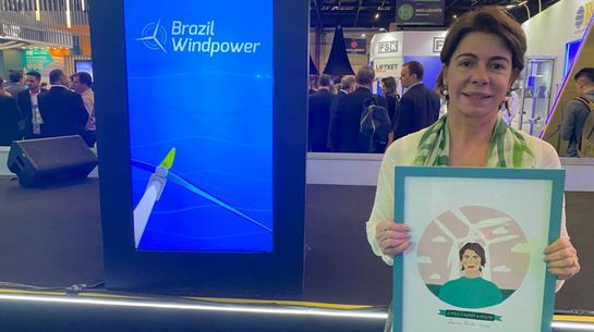 Foto da executiva segurando quadro com ilustração que recebeu como homenagem; ao fundo, placa com as palavras &quot;Brazil Windpower&quot;