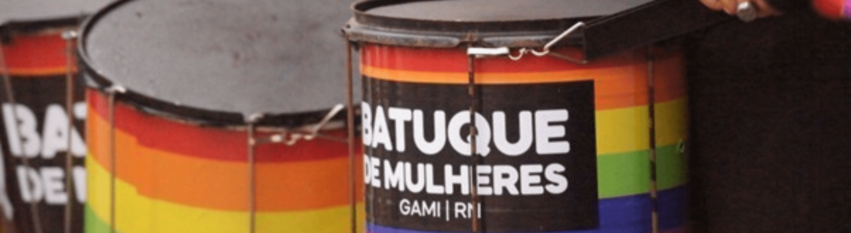Dois instrumentos de percussão pintados nas cores da bandeira LGBTQIAPN+, com adesivos com o fundo preto e escrito "Batuque de mulheres"
