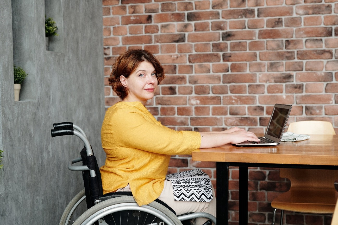 Mulher sentada em uma cadeira de rodas, de frente para uma mesa, digitando em um notebook enquanto olha para a câmera.