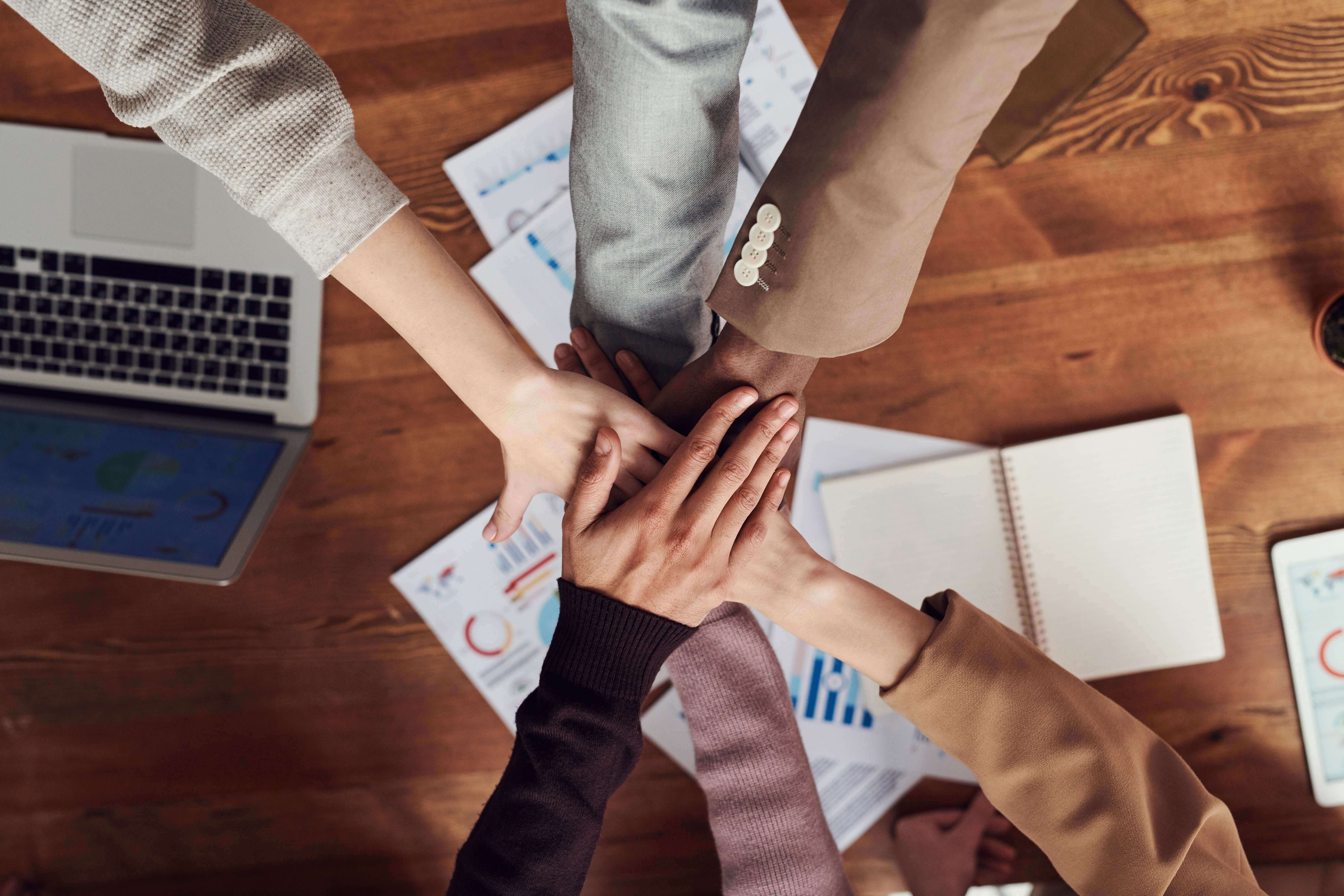Mãos juntas em cima de mesa de madeira com papéis, caderno e notebook ao fundo