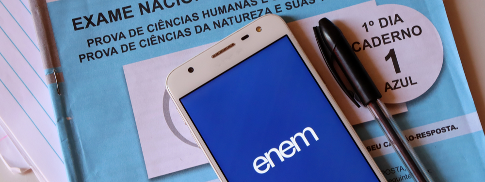 foto de um celular em cima de uma prova do Enem, o celular é branco e a imagem que aparece na tea é de fundo azul e no centro é possível vem o logo do Enem em branco
