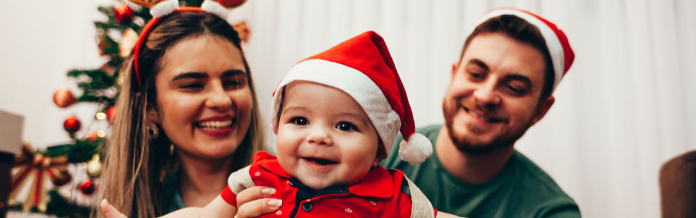foto de uma um casal de homem e mulher com um bebe no colo, eles estão todos sorrindo e ao fundo é possível perceber o clima de natal pelas decorações e colore