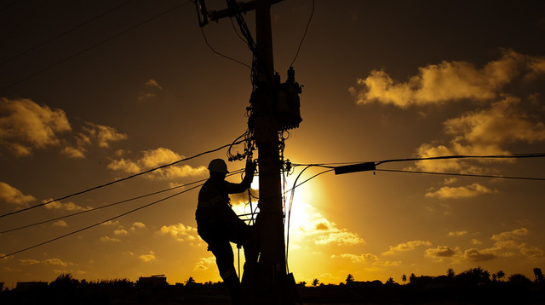 Foto de eletricistas trabalhando em poste, com pôr do sol ao fundo